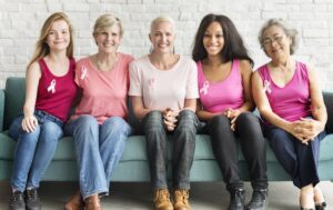 Collection of women sitting together, wearing breast cancer ribbons on their shirts.