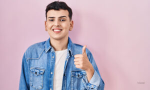 Non binary person standing over pink background smiling happy and positive, thumb up doing excellent and approval sign